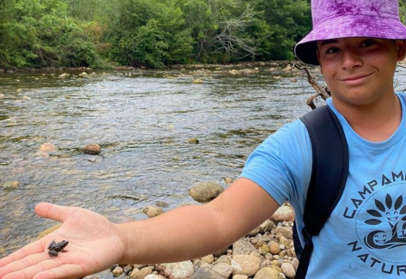 Adolescente con una rana en la mano frente al río con una camisa del campamento multiaventura Naturkids